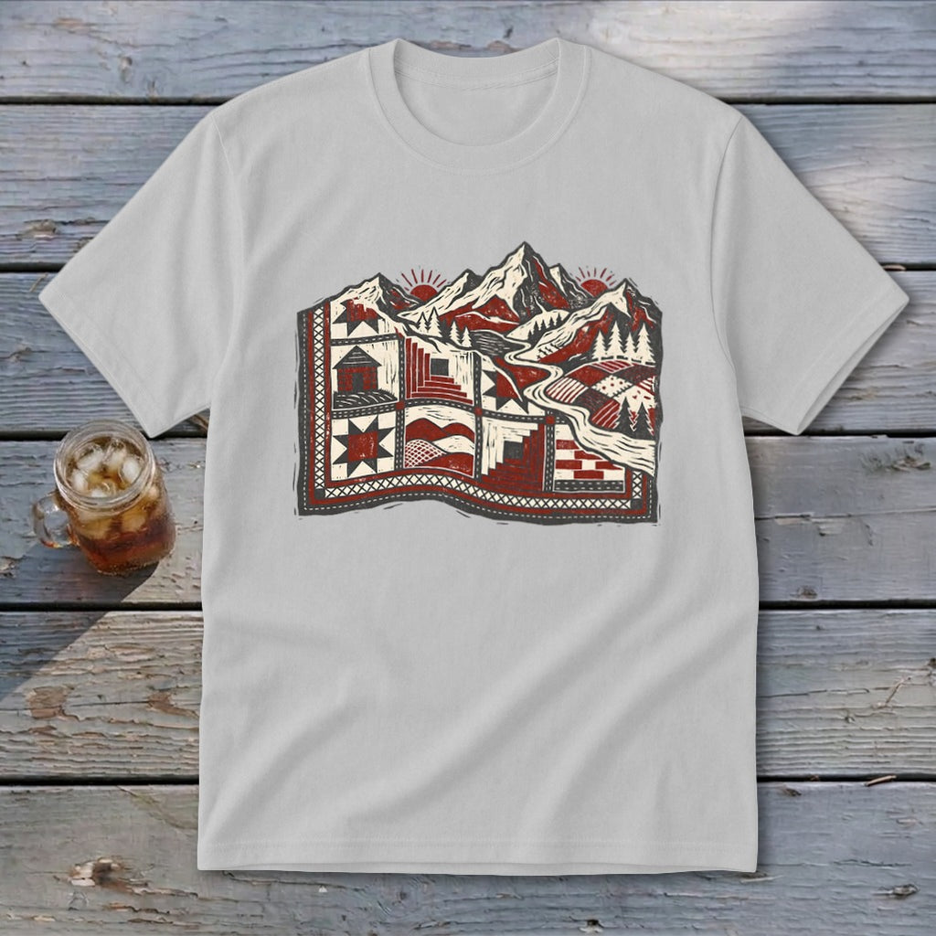 A light grey t-shirt featuring a stylized graphic of a patchwork quilt landscape with mountains, trees, and suns.