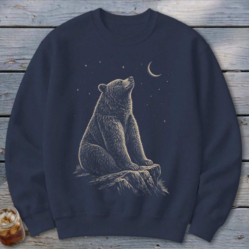 Bear sitting on rock under crescent moon and stars illustration on navy blue sweatshirt.