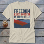 A cream-colored t-shirt with blue and red text reading "Freedom Rings Loudest in These Hills" above a mountain graphic.