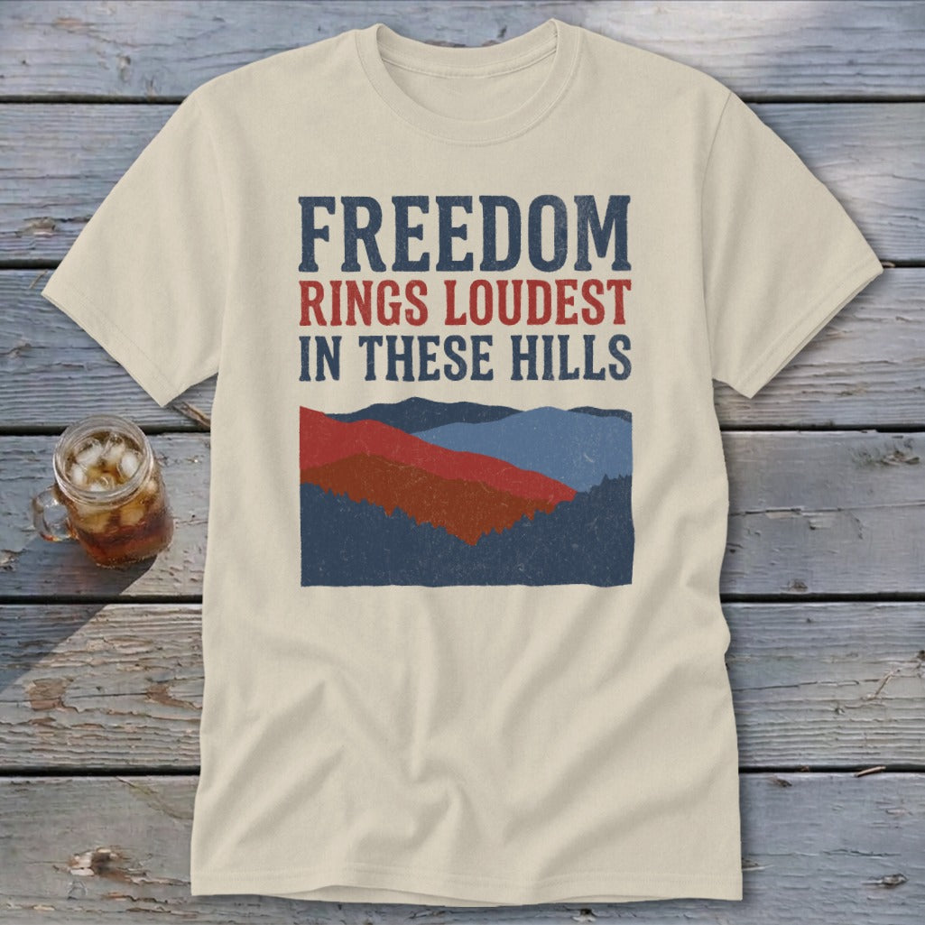 A cream-colored t-shirt with blue and red text reading "Freedom Rings Loudest in These Hills" above a mountain graphic.
