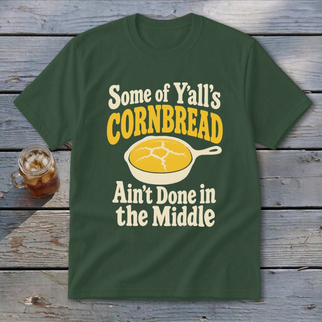 Dark green t-shirt featuring yellow and white text that reads Some of Y'all's Cornbread Ain't Done in the Middle above a skillet of cornbread.