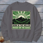 A dark gray crewneck sweatshirt featuring a graphic of green mountains, sunbursts, and the text Born in the Holler Raised on the Ridge.