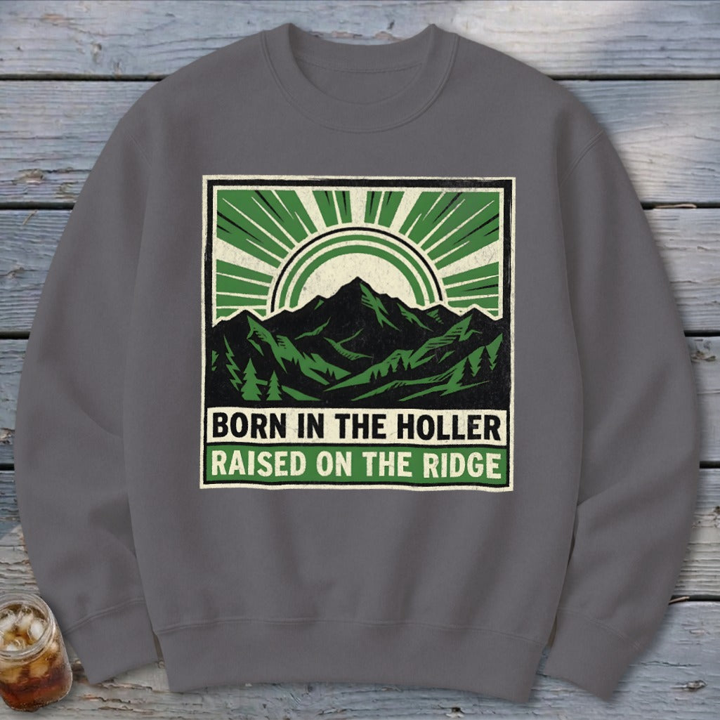 A dark gray crewneck sweatshirt featuring a graphic of green mountains, sunbursts, and the text Born in the Holler Raised on the Ridge.