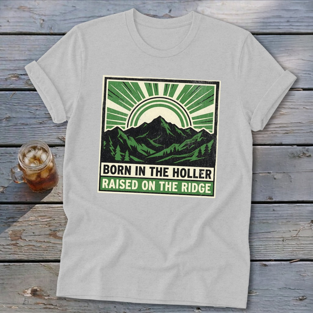 A light gray t-shirt with a square graphic depicting mountains, a sun, and the text BORN IN THE HOLLER RAISED ON THE RIDGE.
