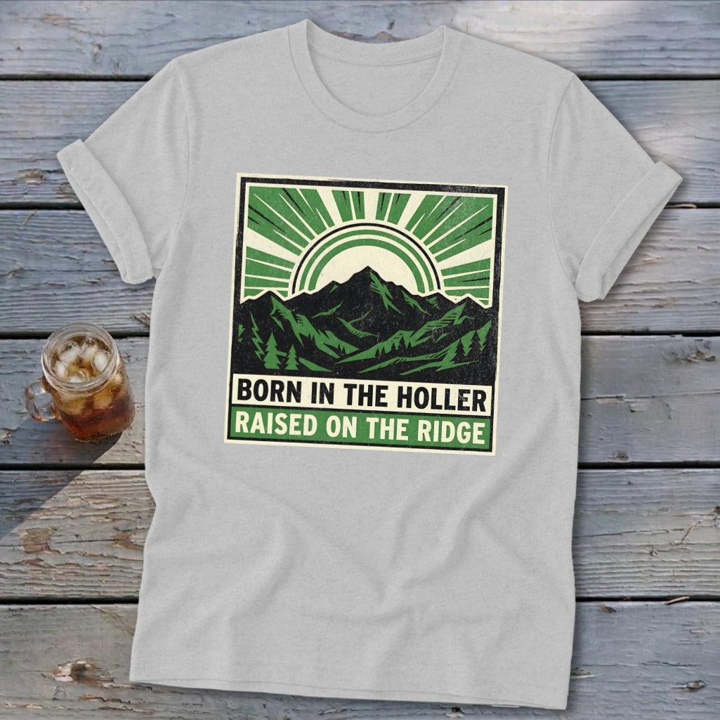 A light gray t-shirt with a square graphic depicting mountains, a sun, and the text BORN IN THE HOLLER RAISED ON THE RIDGE.