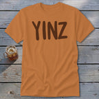 Orange t-shirt with brown text "YINZ" on a wooden background.