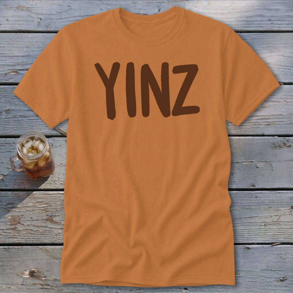 Orange t-shirt with brown text "YINZ" on a wooden background.
