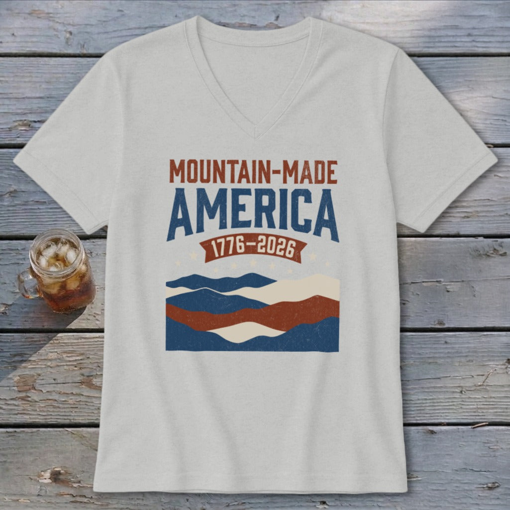 A light gray V-neck t-shirt featuring a retro mountain graphic with the text MOUNTAIN-MADE AMERICA and 1776-2026.