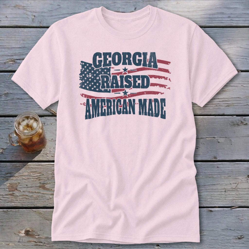 A light pink t-shirt featuring a distressed American flag graphic with the text GEORGIA RAISED AMERICAN MADE.