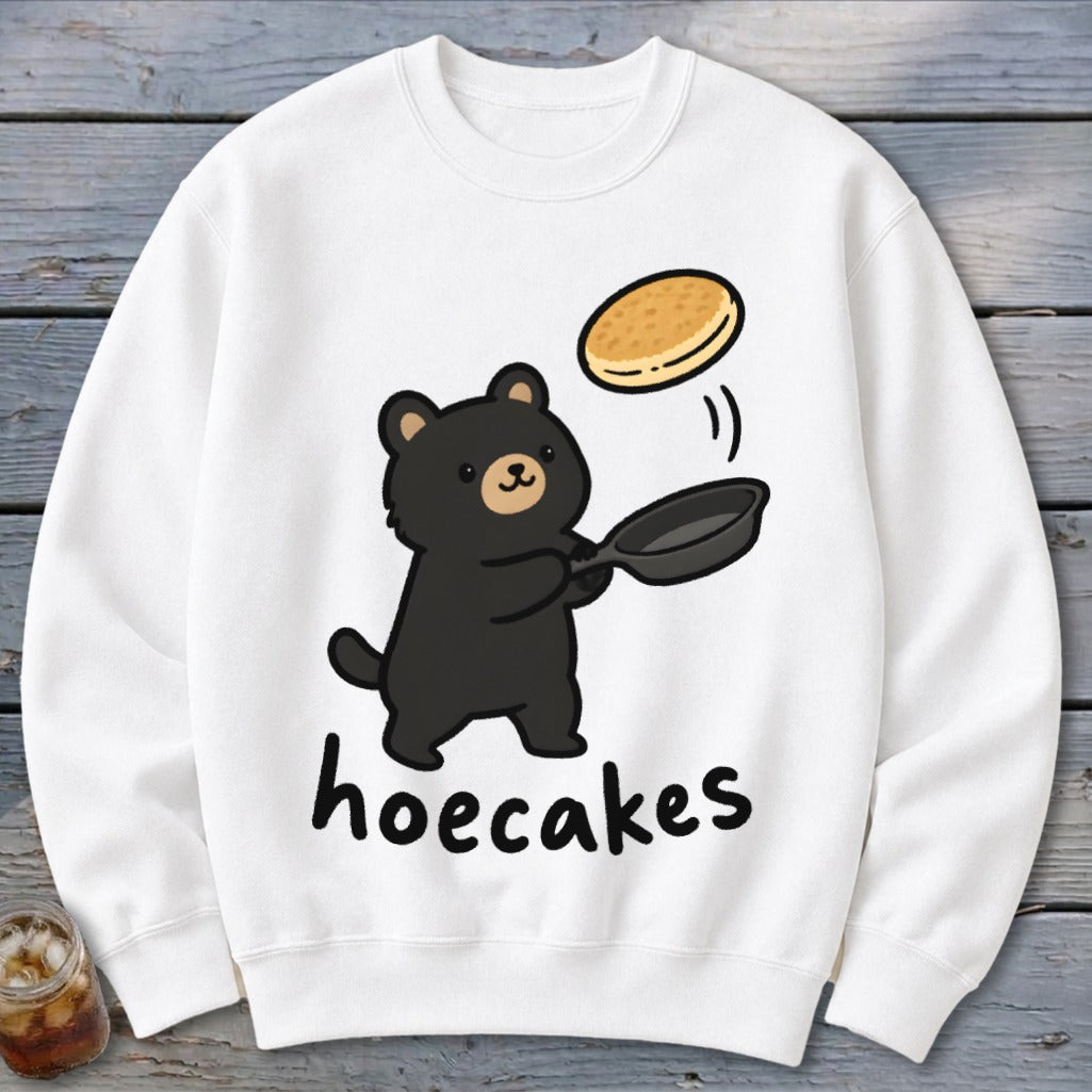 A white crewneck sweatshirt features a cute cartoon black bear flipping a pancake from a skillet, with the text hoecakes underneath.