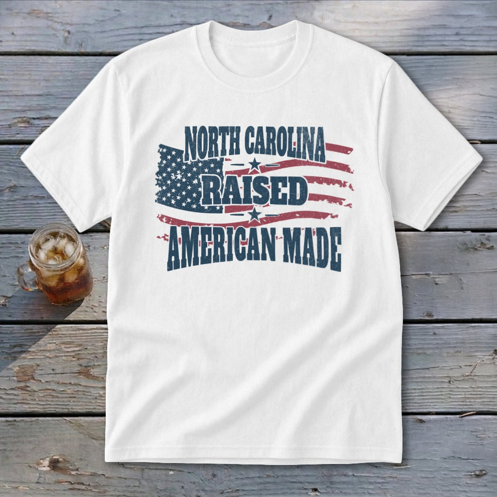 A white short-sleeved t-shirt featuring the text North Carolina Raised American Made over a distressed American flag graphic, lying on a wooden surface beside a glass of iced tea.
