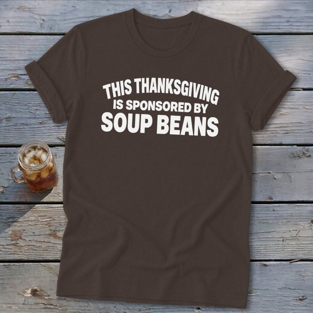 A brown t-shirt featuring the white text "THIS THANKSGIVING IS SPONSORED BY SOUP BEANS" lies on a wooden surface.