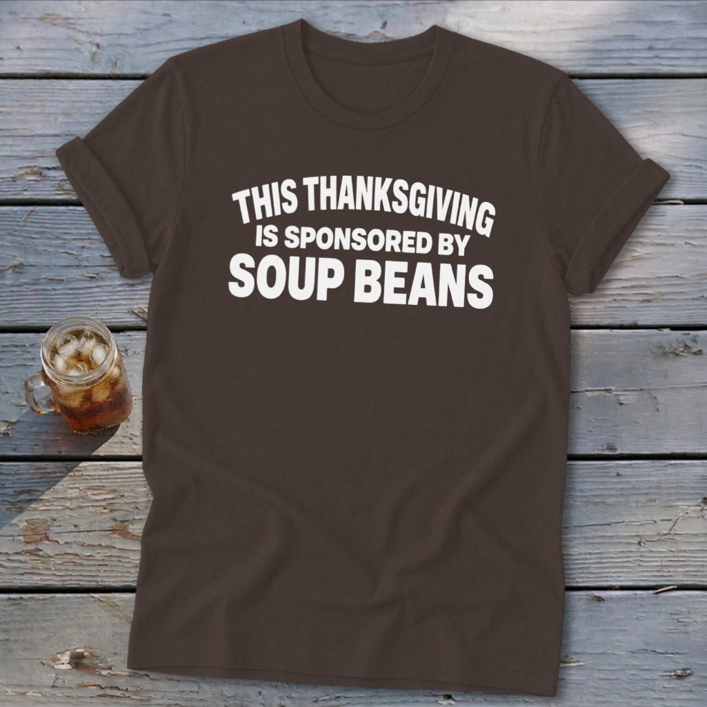 A brown t-shirt featuring the white text "THIS THANKSGIVING IS SPONSORED BY SOUP BEANS" lies on a wooden surface.