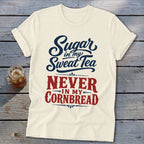 A cream-colored crewneck t-shirt with rolled sleeves features blue and red lettering reading Sugar in my Sweet Tea NEVER IN MY CORNBREAD laid against a rustic wooden background alongside a glass of iced tea.