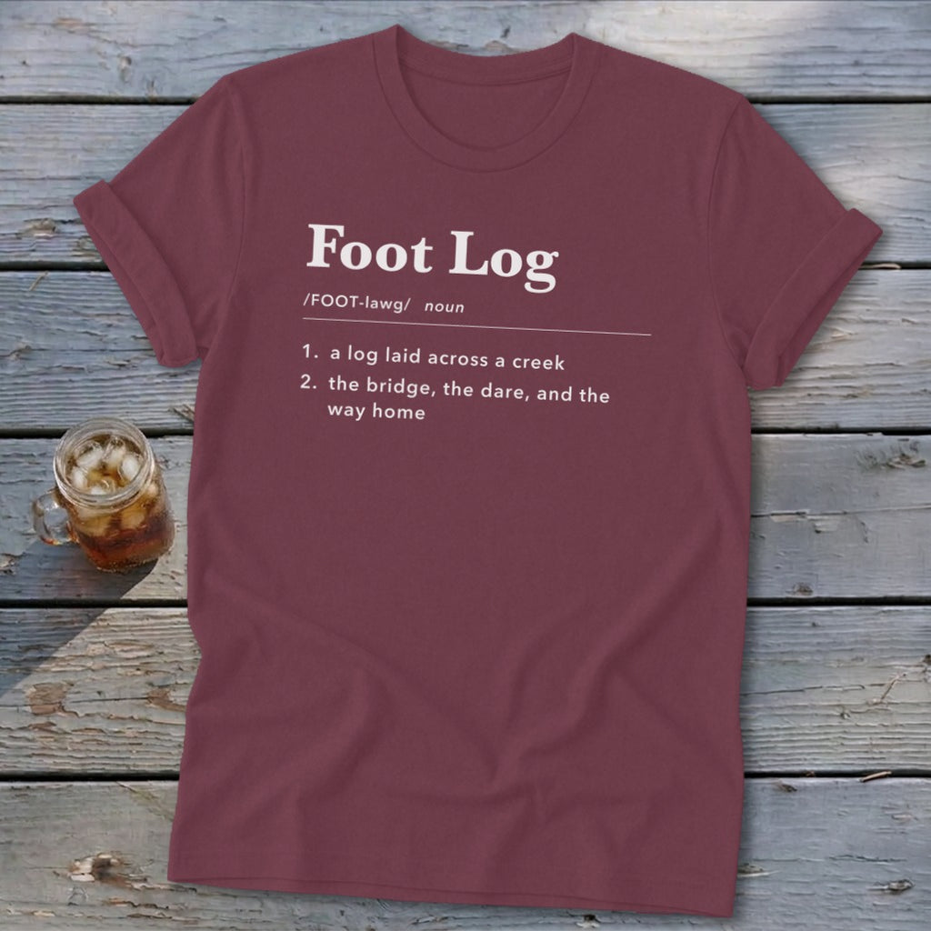 A dark red crewneck t-shirt featuring dictionary-style text that defines a foot log on a wooden background.