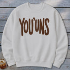 White crewneck sweatshirt with brown "YOU'UNS" text graphic.