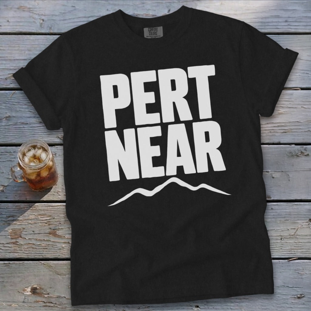 A black t-shirt with white text reading PERT NEAR above a simple mountain outline, resting on a weathered wooden surface.