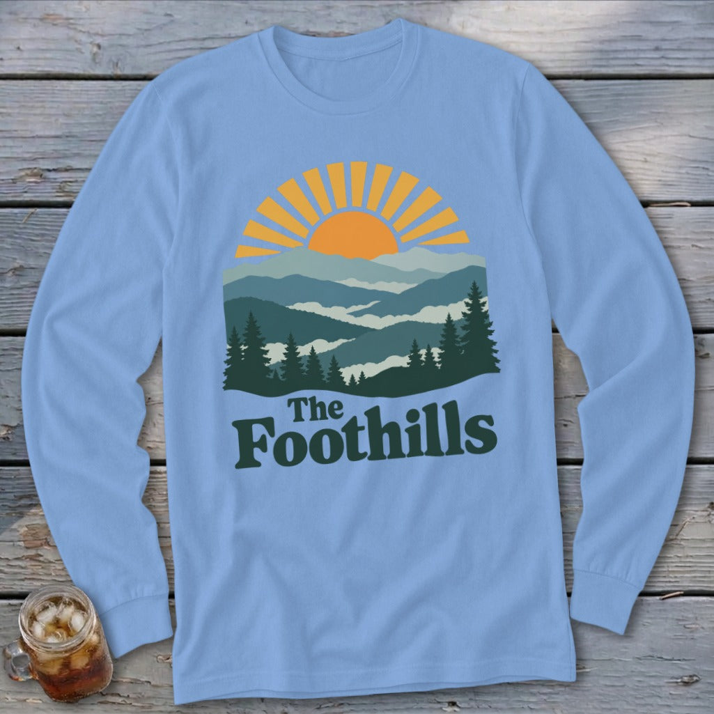 Light blue long sleeve shirt with a mountain and sunrise graphic and "The Foothills" text.