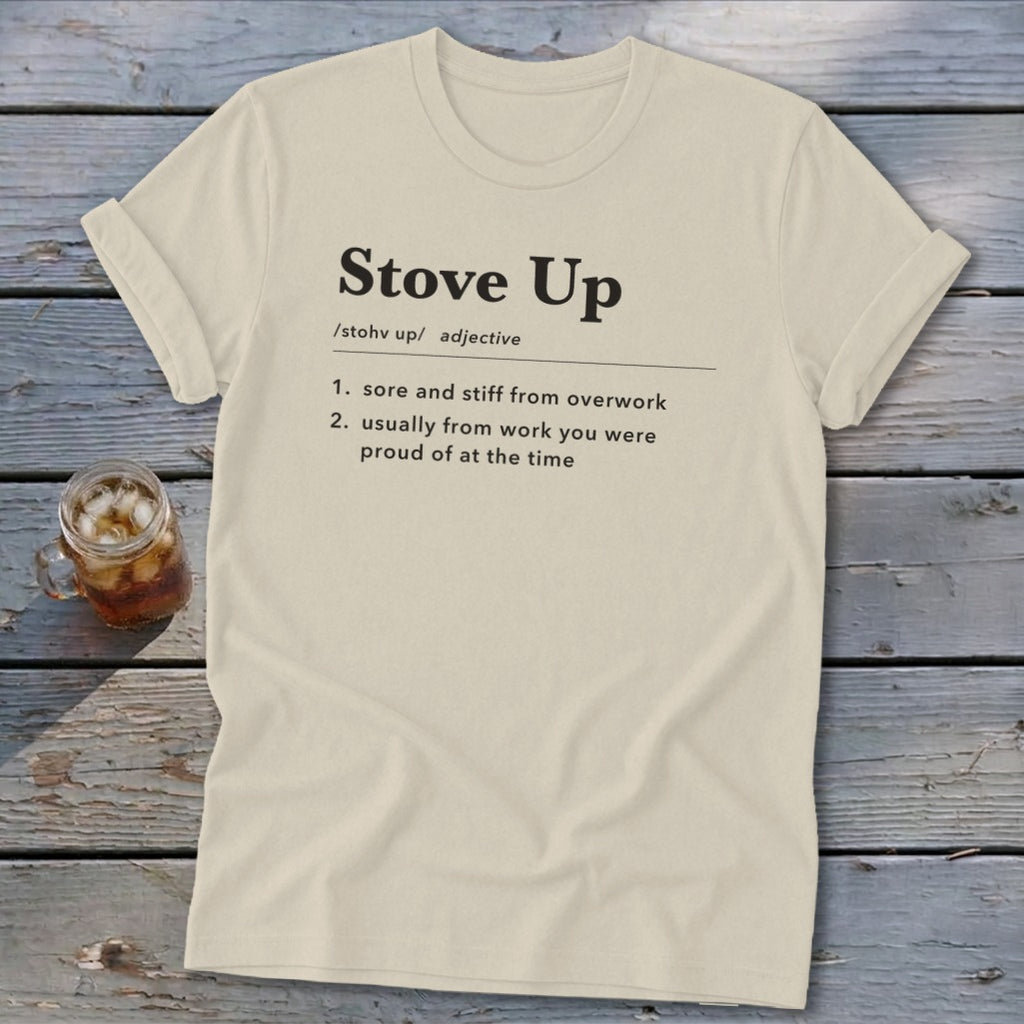 A cream-colored t-shirt with rolled sleeves lies on a rustic wooden table, featuring dictionary-style text defining Stove Up.