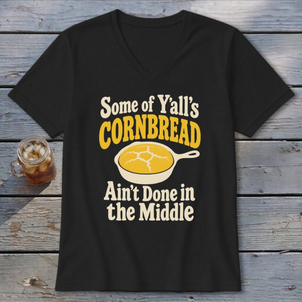 Black V-neck t-shirt with graphic text reading Some of Y’all’s Cornbread Ain’t Done in the Middle surrounding an illustration of cornbread in a skillet.