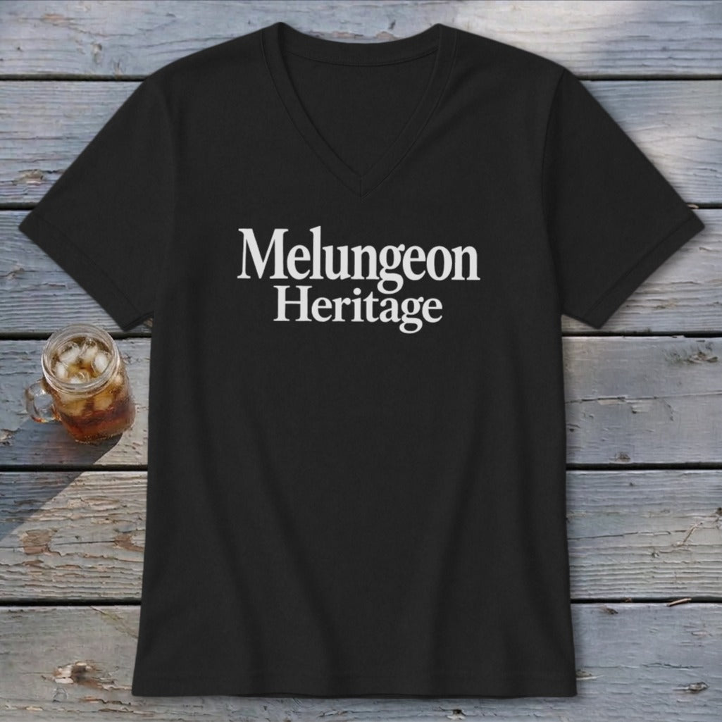 A black V-neck t-shirt with white Melungeon Heritage text lies flat on weathered wooden boards next to an iced beverage.