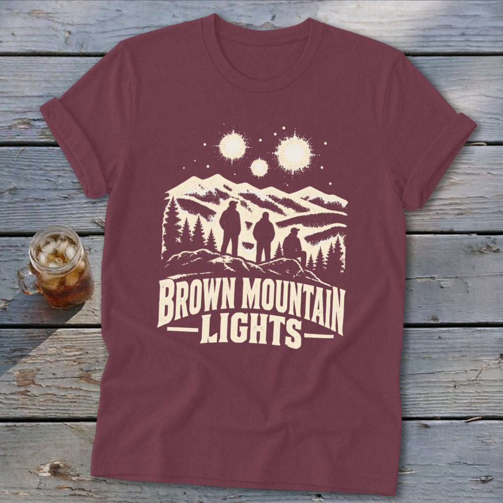 Maroon t-shirt featuring a mountain graphic, three figures, glowing lights above, and the text Brown Mountain Lights.