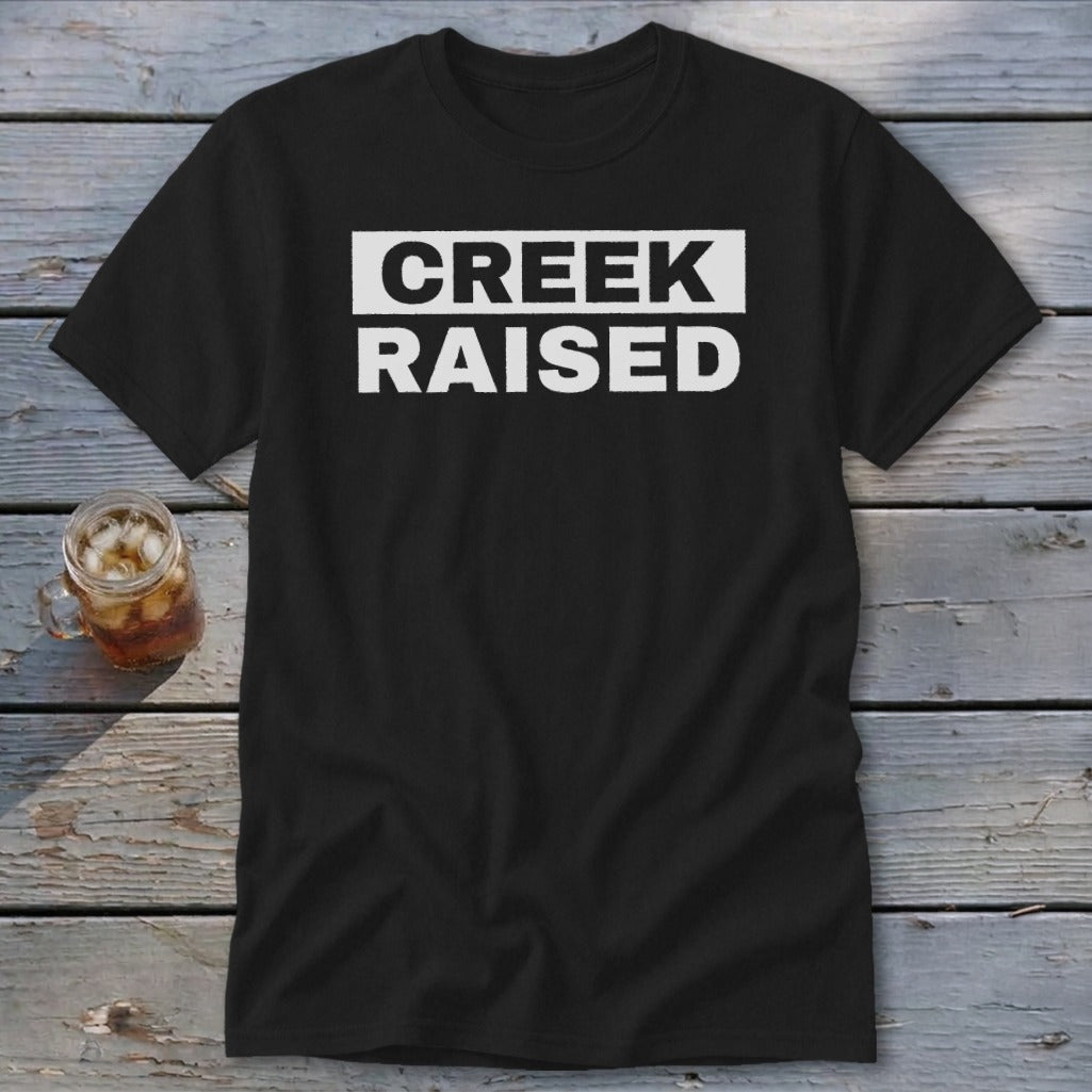 Black t-shirt with white text reading "CREEK RAISED" on a rustic wooden background.