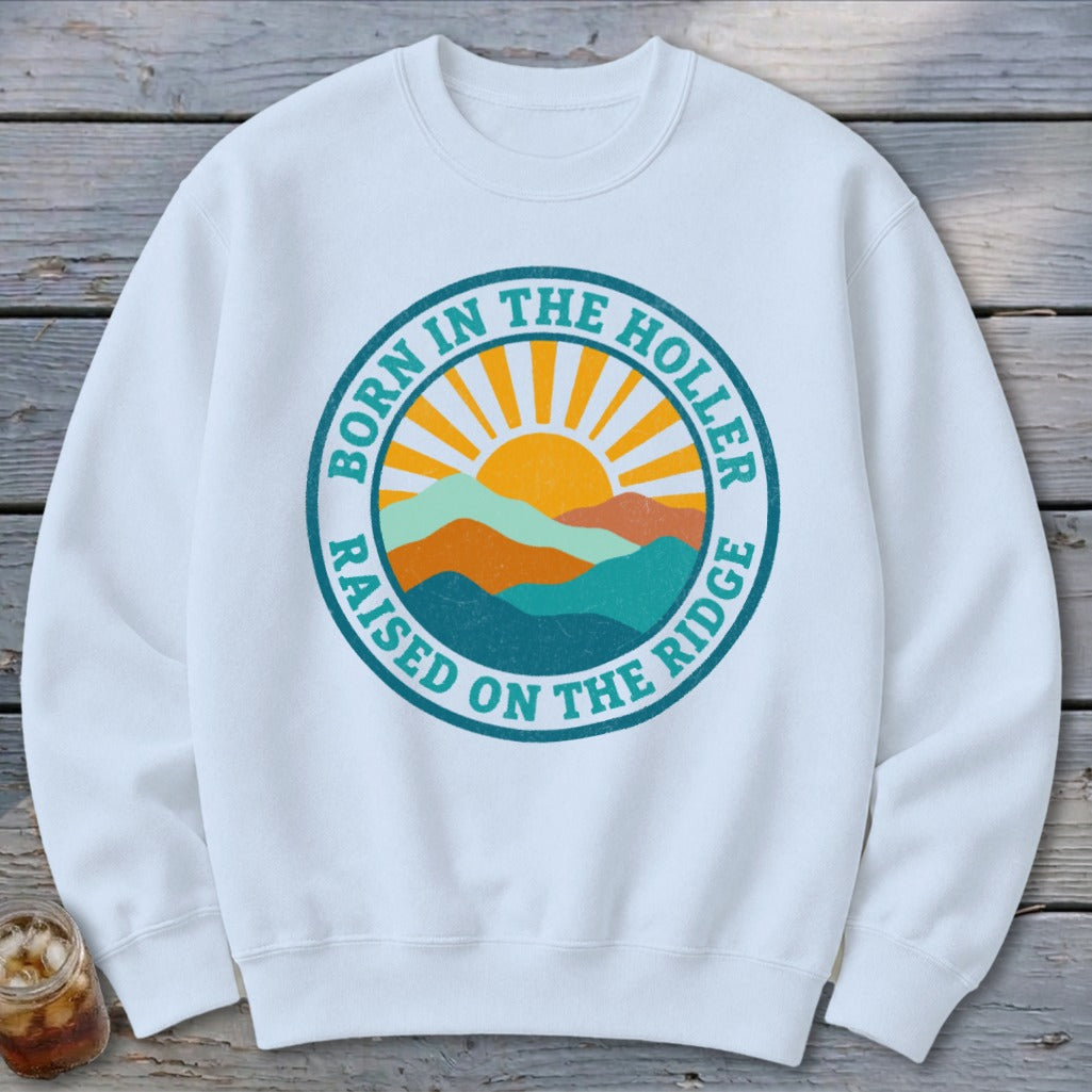 White crewneck sweatshirt featuring a circular graphic with mountains, a sun, and the text Born in the Holler Raised on the Ridge.