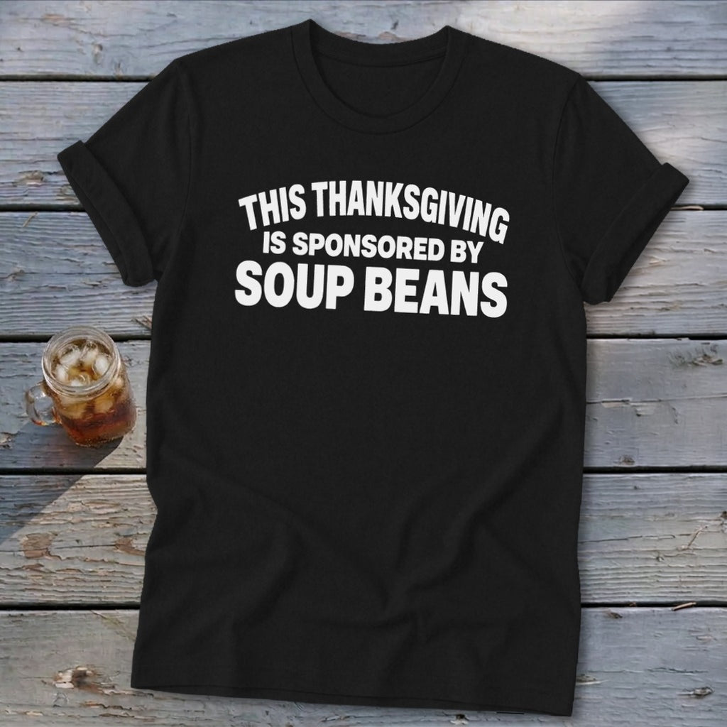 A black t-shirt with white text that reads THIS THANKSGIVING IS SPONSORED BY SOUP BEANS on a wooden background.