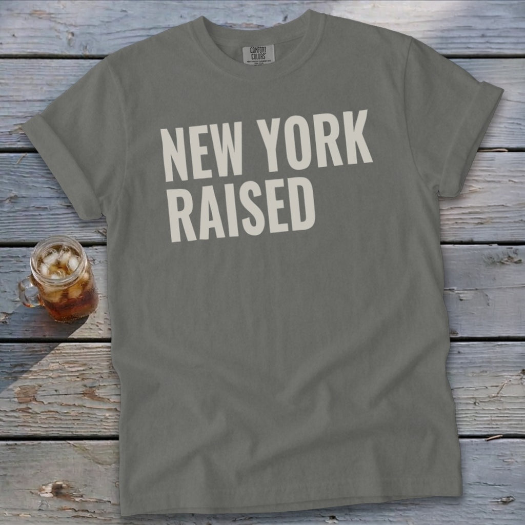 Grey t-shirt with white text reading "NEW YORK RAISED" on a wooden surface.