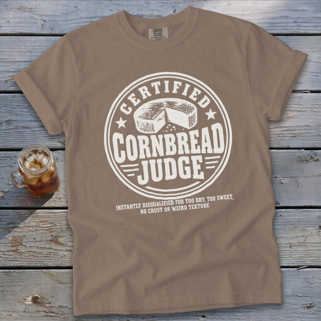A brown t-shirt featuring a circular white graphic with the text "Certified Cornbread Judge."