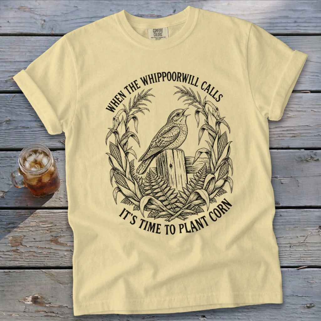A pale yellow t-shirt featuring a black graphic of a bird on a wooden post surrounded by corn and fern fronds, with text reading "WHEN THE WHIPPOORWILL CALLS IT'S TIME TO PLANT CORN".