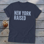Navy t-shirt with light blue "NEW YORK RAISED" text, on a wooden surface.