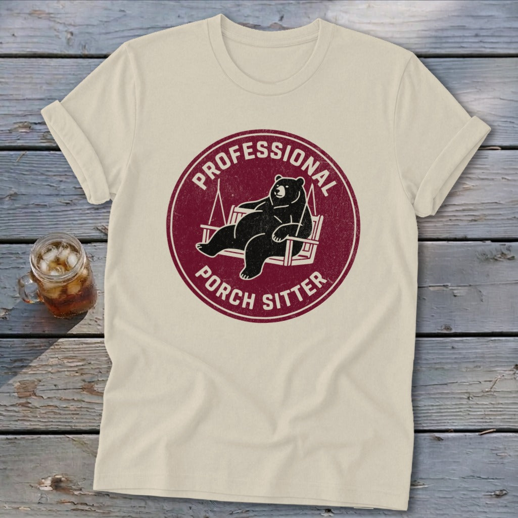 Cream-colored t-shirt featuring a maroon circular graphic of a bear lounging on a porch swing, with text reading Professional Porch Sitter.