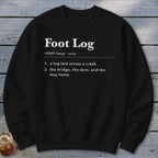 A black crewneck sweatshirt printed with a dictionary definition of Foot Log in white text, laid flat on a rustic wooden background.