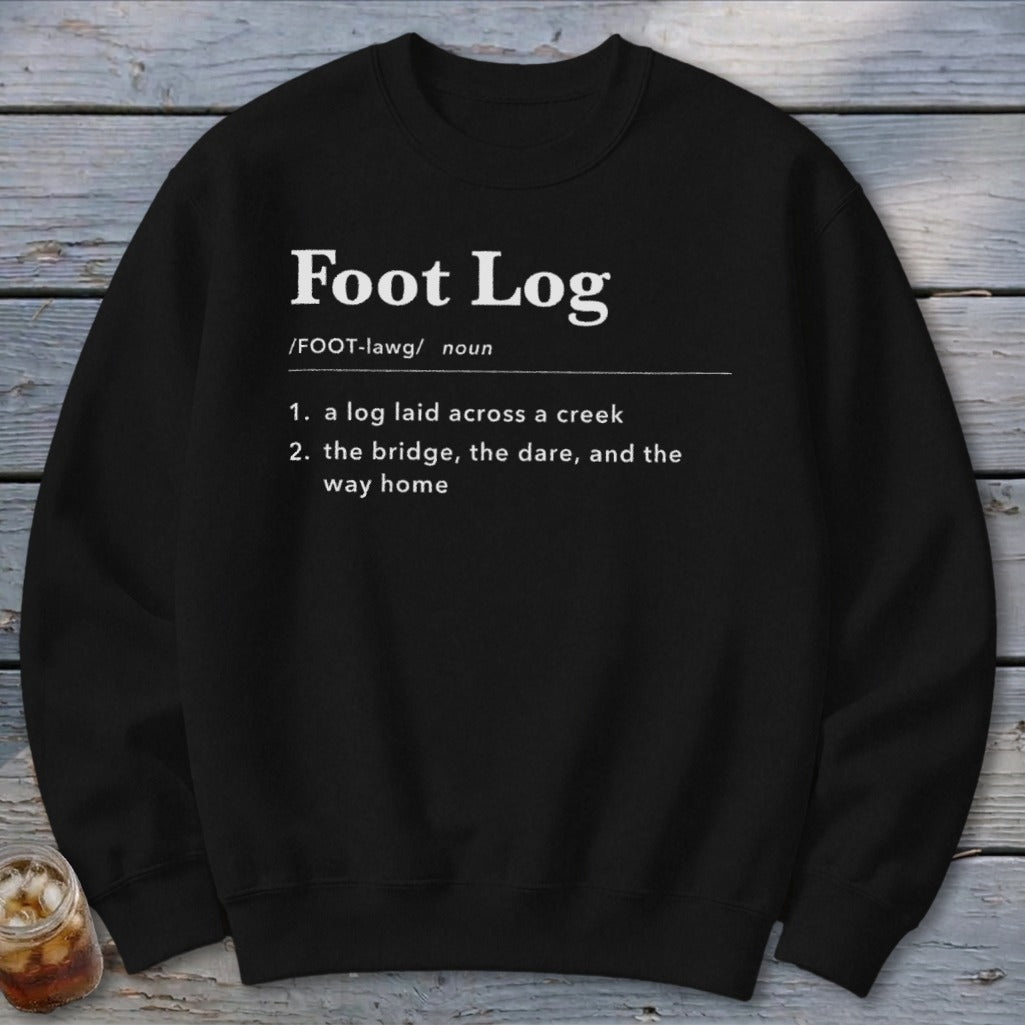 A black crewneck sweatshirt printed with a dictionary definition of Foot Log in white text, laid flat on a rustic wooden background.