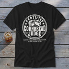 A black t-shirt printed with a white graphic reading Certified Cornbread Judge surrounding a cornbread illustration.