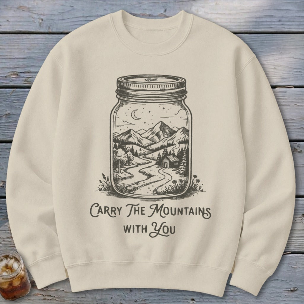 A beige crewneck sweatshirt printed with a mason jar containing a mountain landscape and text reading Carry The Mountains With You.
