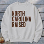 White sweatshirt with brown text reading "NORTH CAROLINA RAISED".