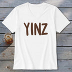 White v-neck t-shirt with brown "YINZ" text and a glass of iced drink.