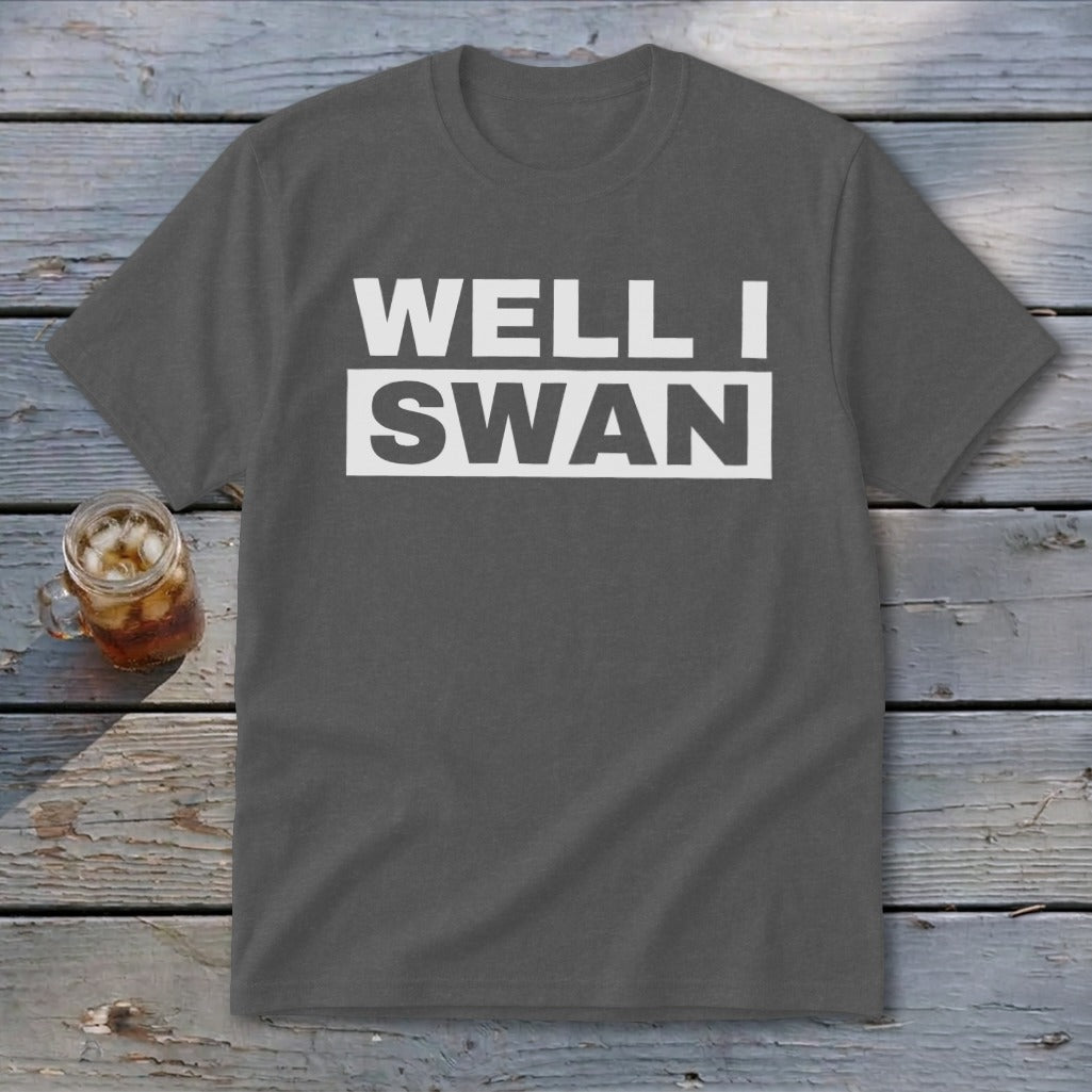Dark gray t-shirt with white block text reading WELL I SWAN laid on a weathered wooden surface.