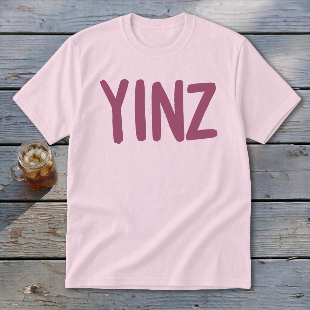 Light pink t-shirt with maroon "YINZ" text on a wooden surface with a drink.
