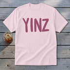 Light pink t-shirt with maroon "YINZ" text on a wooden surface with a drink.
