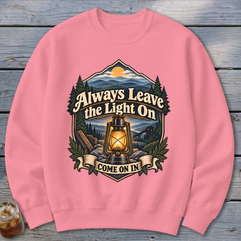 Pink sweatshirt with a graphic of a lantern and mountains, reading "Always Leave the Light On Come On In".