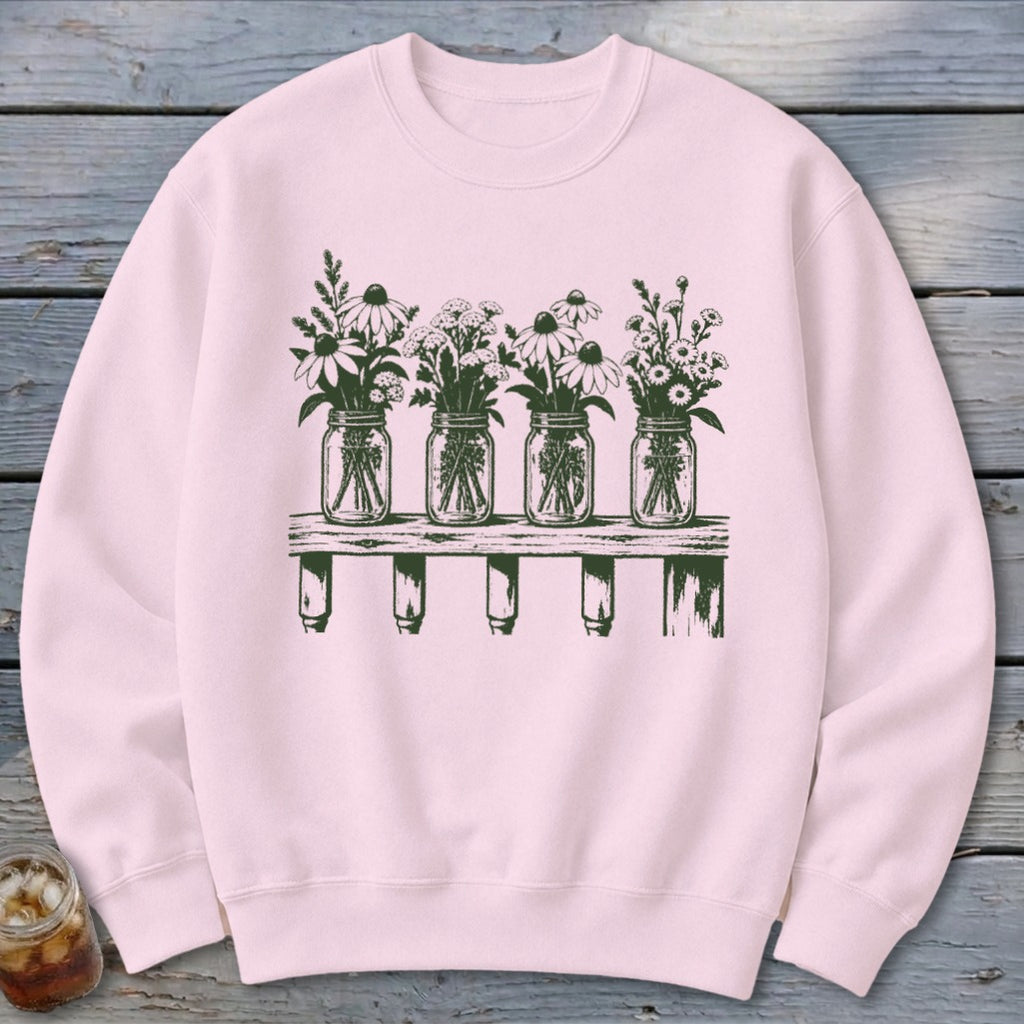 A light pink crewneck sweatshirt featuring a dark green graphic of four mason jars filled with wildflowers on a wooden shelf.