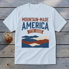 A light blue t-shirt displaying a vintage graphic with red and blue typography reading "Mountain-Made America 1776-2026" above stylized mountains.