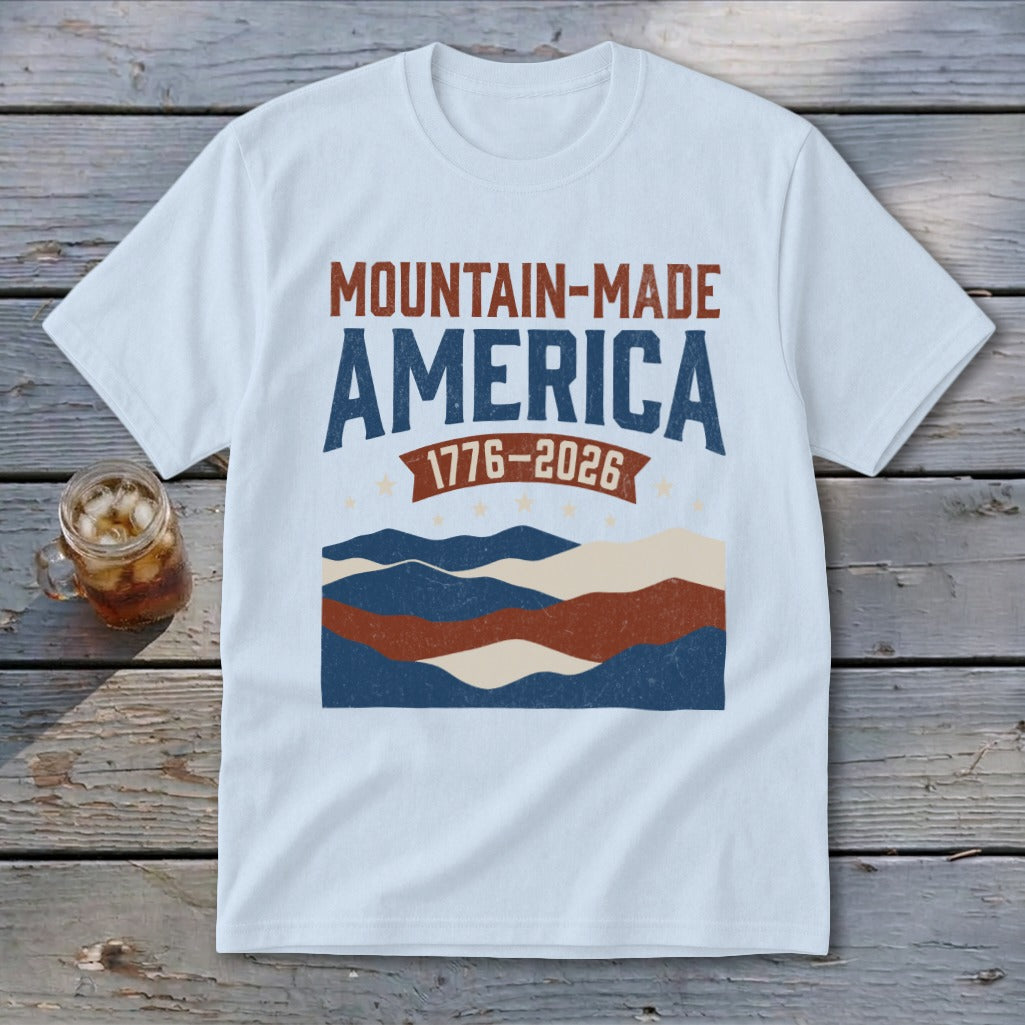 A light blue t-shirt displaying a vintage graphic with red and blue typography reading "Mountain-Made America 1776-2026" above stylized mountains.