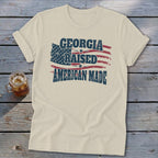 Beige short-sleeve t-shirt with rolled cuffs, featuring a distressed American flag and text that reads GEORGIA RAISED AMERICAN MADE.