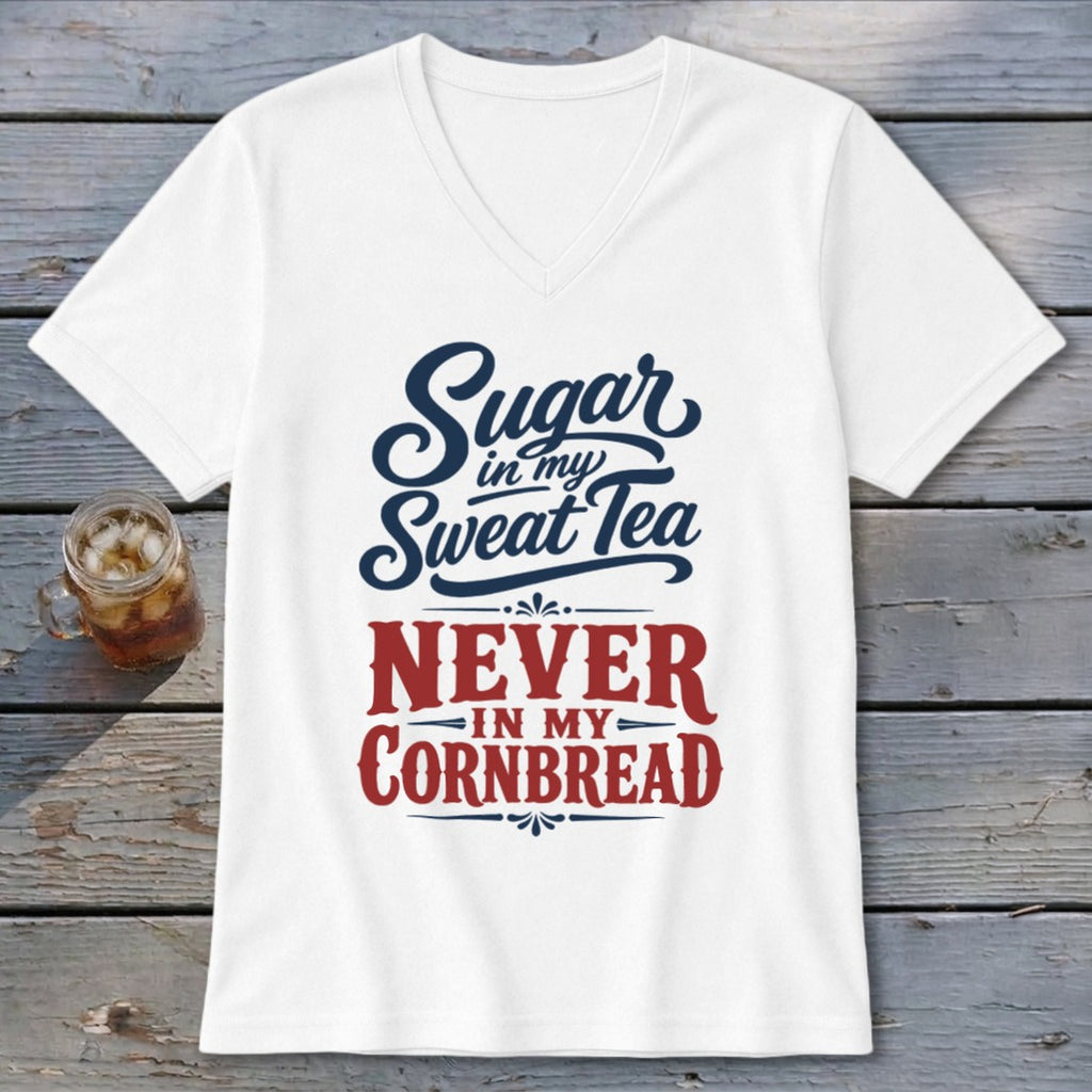 A white V-neck t-shirt with blue and red text reading Sugar in my Sweet Tea NEVER IN MY CORNBREAD rests on a rustic wooden surface next to a glass of iced tea.