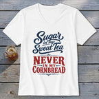 A white V-neck t-shirt with blue and red text reading Sugar in my Sweet Tea NEVER IN MY CORNBREAD rests on a rustic wooden surface next to a glass of iced tea.